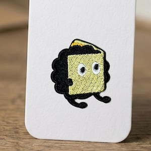 May include: Embroidered patch showing a cartoon slice of cake. The cake has a yellow body, black frosting, and two white-eyed, black-pupiled eyes. It has a black, wavy border and small black feet. The cake slice has a small, gold-coloured top.