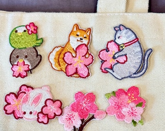 May include: Embroidered patches featuring a green bird, a brown penguin, a Shiba Inu dog, a gray cat, and a white bunny, each holding or surrounded by pink cherry blossoms. Additional patches include cherry blossom sprigs. The patches are on a cream-colored fabric.