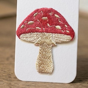 May include: Embroidered mushroom patch with a red cap and tan stem. The mushroom design is detailed with tan stitching on the cap and stem, set against a white background. This patch is suitable for clothing or accessories.