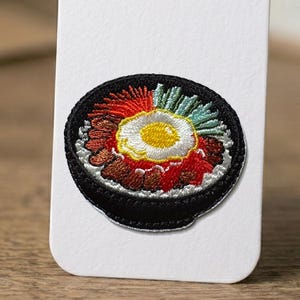 May include: Embroidered patch featuring a colorful bowl of food with a fried egg in the center. The patch has a black border and includes red, brown, green, and yellow thread. The design is likely inspired by a Korean dish.