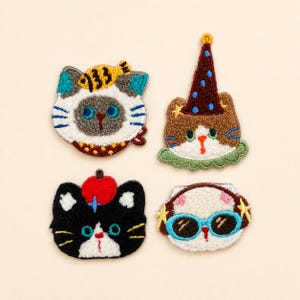 May include: Four embroidered cat patches featuring distinct designs: a cat with a yellow fish on its head, a brown cat wearing a blue-spotted party hat, a black cat with a red apple on its head, and a white cat wearing blue-rimmed sunglasses.