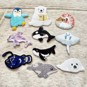 May include: A collection of embroidered patches depicting marine life, such as a penguin, polar bear, orca, dolphin, stingray, and anglerfish. The patches are colourful and detailed, with a textured finish. These patches are designed for decorative use.