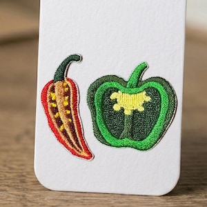 May include: Embroidered patches of a red chili pepper and a green bell pepper. The chili pepper is red with yellow and brown details, while the bell pepper is green with a yellow center. Both patches have green stems and are on a white background.