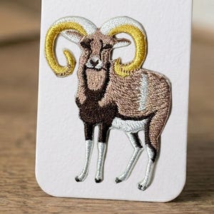 May include: Embroidered patch featuring a ram with large, curved golden horns. The ram has a brown body, dark brown face and legs, and white accents. The patch is likely for clothing or accessories.