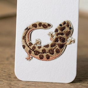 May include: Embroidered patch featuring a gecko in shades of brown and tan with darker brown spots. The gecko is curled in a relaxed pose, with its tiny feet and tail visible. The patch is likely designed to be ironed or sewn onto fabric.