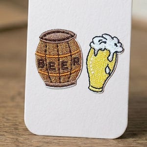 May include: Embroidered patches of a beer barrel and a pint of beer. The barrel is brown with the word "BEER" stitched across it. The beer glass is yellow with white foam. The patches are on a white background.