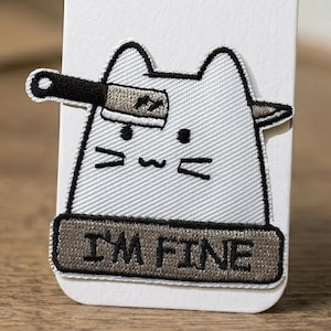 Könnte beinhalten: Gestickter Aufnäher mit einer Cartoon-Katze mit einem Messer. Die Katze ist hellgrau mit schwarzen Umrissen. Der Aufnäher hat den Text "I'M FINE" in Dunkelbraun. Ein humorvolles Design.