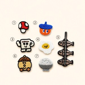 May include: A collection of seven embroidered patches featuring various designs: a red and white mushroom, a blue and orange acorn, a white mug with a face, a fried egg, three brown fish on a string, a tan acorn character, and a bowl of white rice.