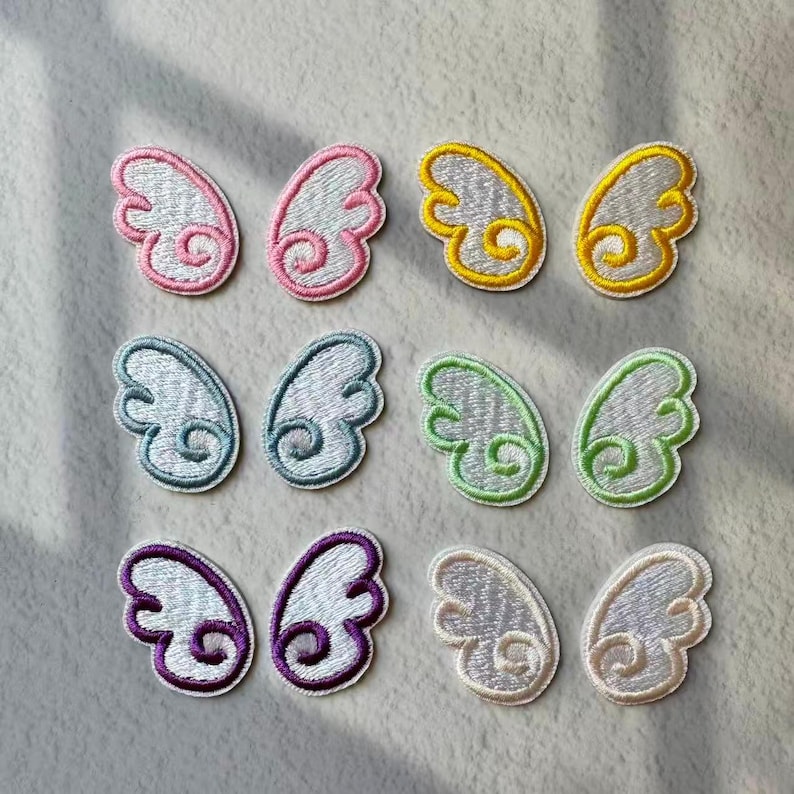 May include: A collection of embroidered wing patches in various colors. The patches feature a white base with colorful outlines in pink, yellow, blue, green, purple, and cream. Each patch is approximately 1.5 inches wide and 1 inch tall.