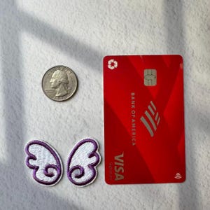 May include: A red Bank of America Visa credit card, a US quarter, and two embroidered butterfly wing patches with white and purple details are arranged on a white surface. The card has the Visa Signature logo.