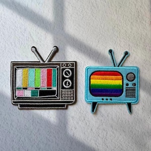 May include: Two embroidered patches of vintage televisions. One is black with a colorful test pattern, the other is light blue with a rainbow design. Both have antennas and are on a white background.