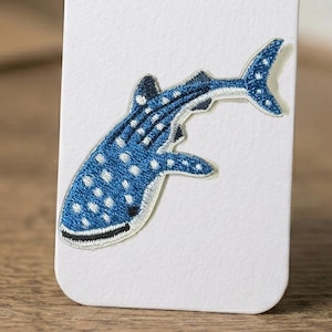 Könnte beinhalten: Gestickter Aufnäher mit einem blauen Walhai mit weißen Punkten. Der Aufnäher ist mit schwarzen Nähten an den Rändern und am Maul versehen. Der Aufnäher befindet sich auf weißem Hintergrund.