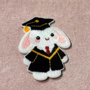 Op de afbeelding: Geborduurde patch van een witte konijn met een zwarte afstudeerhoed en toga. Het konijn heeft roze oren en wangen, een rode stropdas en gouden randen op de toga. De patch is ontworpen in een schattige, cartoonachtige stijl.