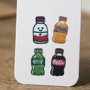 May include: Four embroidered patches featuring miniature beverage bottles. The top left is white with red and green stripes, the top right is orange with blue text, the bottom left is green with white text, and the bottom right is black with red text.