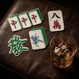 May include: A collection of embroidered Mahjong patches and a glass of whiskey with ice cubes on a brown leather surface. The patches feature Chinese characters and colorful designs, with green and red accents.