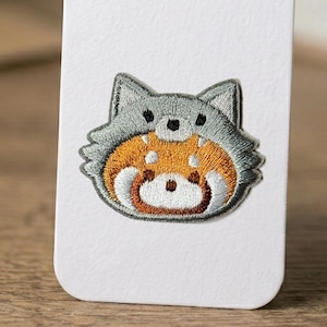 May include: Embroidered patch featuring a red panda wearing a gray wolf hat. The patch has a detailed design with orange, white, and brown fur, and gray accents. The patch is likely for clothing or accessories.