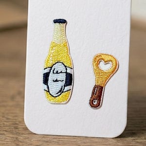 以下が含まれることがあります： 青と白のラベルが付いた黄色のビール瓶と、金の栓抜きが刺繍されたパッチセット。栓抜きにはハート型の切り抜きがあります。パッチは白い背景にあります。
