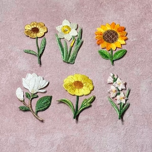 May include: Embroidered floral patches in various designs, including a sunflower, daffodil, and other flowers. Each patch features detailed stitching with green leaves and stems, set against a pink background.