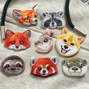 Może przedstawiać: Haftowane naszywki przedstawiające różne zwierzęta, w tym lisa, szopa pracza, królika, Shiba Inu, leniwca, pandę rudą i wydrę. Każda naszywka prezentuje szczegółowe szwy i żywe kolory, idealne do personalizacji odzieży.