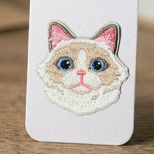 May include: Embroidered patch featuring a cat's face with blue eyes, pink ears, and a beige and white coat. The patch is on a white background. The cat's face is detailed with embroidered thread, showcasing its features.