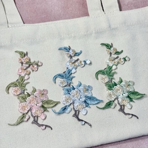 May include: Close-up of a beige tote bag featuring three embroidered floral designs. The designs showcase blossoms in pink, blue, and white, with green leaves and brown stems. The embroidery adds a textured, handcrafted look to the bag.