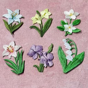 Puede incluir: Parches florales bordados en varios colores, incluyendo blanco, azul claro, amarillo y morado. Cada parche presenta detalles de flores bordadas con hojas y tallos verdes. Los parches están dispuestos sobre un fondo rosa.