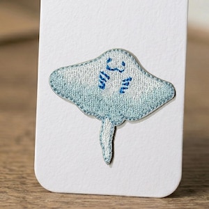 May include: A light blue embroidered stingray patch with a detailed design. The patch features a light blue body with darker blue accents and a detailed outline. The stingray has a tail and is on a white background.