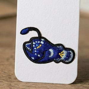 May include: Embroidered patch featuring a deep blue anglerfish with a black outline. The patch has white and yellow accents, and a black stem. The patch is on a white background.