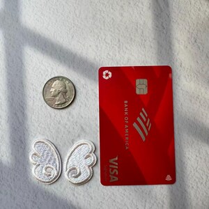 May include: A red Bank of America Visa credit card with the Visa logo and a silver quarter next to two white embroidered butterfly wing patches.
