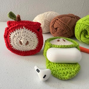 May include: Two crocheted apple-shaped cases for Airpods. One is red and white, the other is green. Both have a simple face design. The cases are sitting on a white surface with balls of yarn in the background.