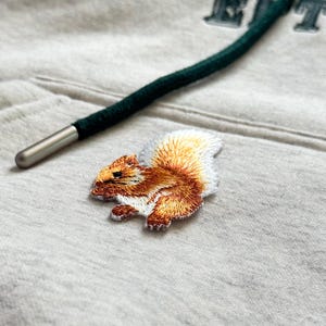 May include: Embroidered squirrel patch in shades of brown, orange, and white, with detailed stitching. The patch is on a light grey fabric, near a dark green drawstring with a silver metal tip. The overall theme is nature and wildlife.