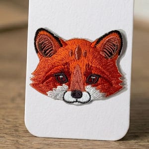 Könnte beinhalten: Gestickter Aufnäher mit einem Fuchskopf. Der Fuchs hat orangefarbenes Fell mit weißen Akzenten um die Schnauze und eine schwarze Nase. Die Augen sind dunkel, und die Ohren haben detaillierte Stickereien. Für Kleidung oder Accessoires.