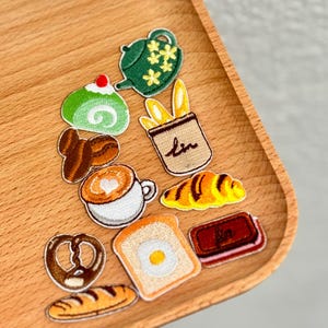 Może przedstawiać: Kolekcja haftowanych naszywek przedstawiających śniadanie i wypieki. Zawiera zielony imbryk, kawałek ciasta, filiżankę kawy, rogalik, chleb i precel. Naszywki znajdują się na drewnianej tacy.