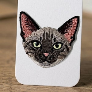 May include: Embroidered cat patch featuring a detailed cat face with green eyes, pink nose, and inner ear detail. The cat's fur is a mix of gray and black, with striped patterns. The patch is likely for clothing or accessories.