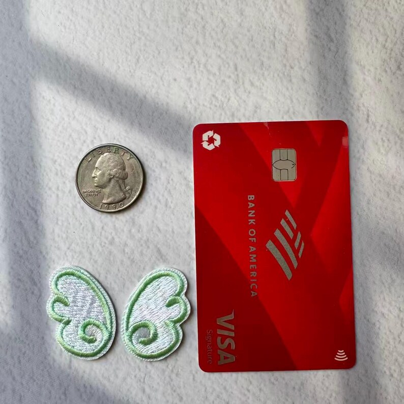 May include: A red Bank of America Visa credit card, a quarter, and two embroidered butterfly wing patches with white and green details are displayed on a white surface.