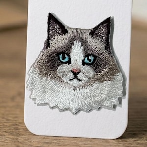 May include: Embroidered patch featuring a cat's face. The cat has gray and white fur, with striking blue eyes and a pink nose. The patch is on a white background, showcasing the detailed stitching and design.
