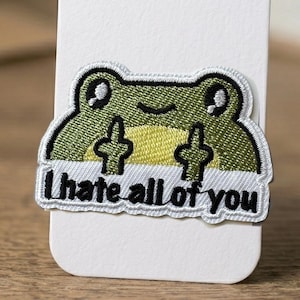 Könnte beinhalten: Gestickter Aufnäher mit einer grünen Frosch-Darstellung mit schwarzen Umrissen und dem Text "I hate all of you". Der Frosch zeigt mit beiden Händen den Mittelfinger. Der Aufnäher hat einen weißen Rand.