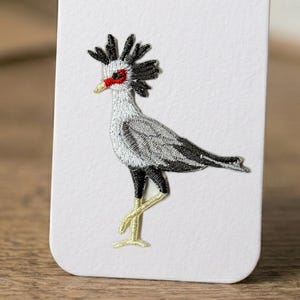 May include: Embroidered patch featuring a Secretary Bird. The bird has a gray body, black wings and legs, and a red face. The patch is on a white background.