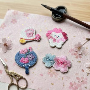 May include: Embroidered patches featuring floral and animal designs. The patches include a white and pink paw print, a pink and white animal, a blue fan with pink flowers, and two flower patches in pink and blue. A paintbrush and inkwell are in the background.