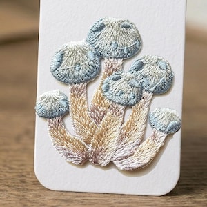 Puede incluir: Diseño bordado de un grupo de setas. Los sombreros de las setas son de color azul claro y blanco, con costuras detalladas. Los tallos son una mezcla de beige y morado claro. El diseño está sobre un fondo blanco.