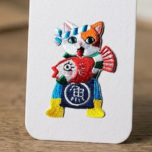 May include: Embroidered patch featuring a cat wearing a blue and white headband and a green scarf, holding a red fish. The cat has orange and white fur and is wearing a blue apron with a Japanese character. Yellow boots complete the design.