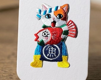 Peut inclure: Écusson brodé représentant un chat portant un bandeau bleu et blanc et une écharpe verte, tenant un poisson rouge. Le chat a des poils orange et blancs et porte un tablier bleu avec un caractère japonais. Des bottes jaunes complètent le design.