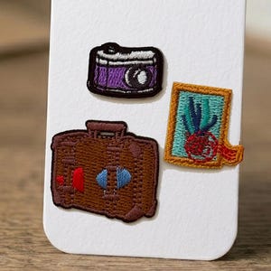 Peut inclure: Trois écussons brodés comprenant un appareil photo violet aux contours noirs, une valise marron avec des accents rouges et bleus, et un cadre rectangulaire doré contenant un motif abstrait bleu sarcelle et rouge sur un support blanc.