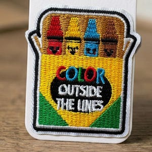 Puede incluir: Parche bordado con una caja de crayones en rojo, amarillo, azul y marrón. El parche tiene las palabras "COLOR OUTSIDE THE LINES" en un óvalo negro sobre un fondo amarillo, con bordes verdes y negros.