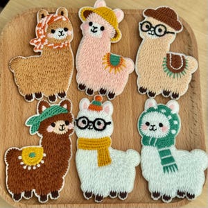 Peut inclure: Six écussons brodés en forme de lama. Chaque lama porte un chapeau, une écharpe ou des lunettes. Les lamas sont présentés sur un plateau en bois, mettant en valeur leurs motifs et couleurs uniques. Idéal pour la couture.