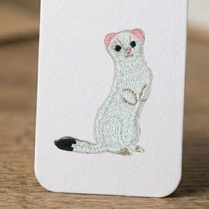 May include: Embroidered white weasel design on a white card. The weasel has pink ears, black eyes, and a black-tipped tail. The weasel is standing upright with its arms crossed. The embroidery is detailed and textured.