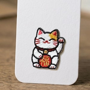 May include: An embroidered patch featuring a white cat with a gold patch on its head, red ears, and whiskers. The cat has one raised paw and holds a round red coin with a gold Chinese character in the center, outlined in black thread.
