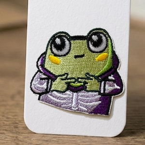 Può includere: Toppa ricamata raffigurante un simpatico ranocchio con una felpa con cappuccio viola. La rana è verde con grandi occhi, macchie gialle sulle guance e un design cucito. La toppa è su sfondo bianco.