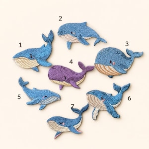 May include: Seven embroidered whale patches arranged in a circular pattern. Six whales are blue with beige underbellies, and one central whale is purple with a beige underbelly. Each patch is numbered from one to seven in black text.