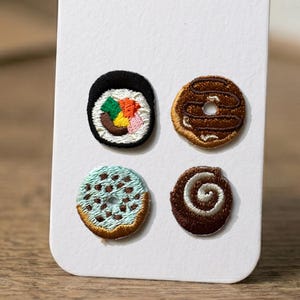 May include: Four embroidered patches featuring food items. There is a sushi roll with colorful fillings, a chocolate donut with a hole, a blue donut with chocolate chips, and a cinnamon roll with a swirl.